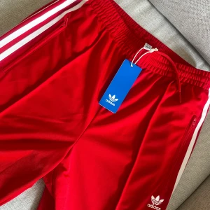 Röda Adidas träningsbyxor med vita ränder - Snygga röda Adidas träningsbyxor med klassiska vita ränder längs sidorna och dragkedjor vid fickorna. Byxorna har resår i midjan med snörning och Adidas-logga på låret. Perfekta för en sportig look.