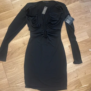 Svart långärmad klänning med cut out från Nelly  - Supersnygg svart långärmad klänning från Nelly med cut out-detaljer och rynkade partier framtill vid brösten. Klänningen har en tight passform. Materialet känns mjukt och stretchigt. Aldrig använd och med prislapp kvar!