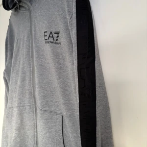 Grå hoodiejacka från EA7 Emporio Armani - Snygg grå hoodiejacka från EA7 Emporio Armani med svarta detaljer längs ärmarna och dragkedja framtill. Jackan har huva, två fickor och logga på bröstet. Perfekt för en sportig och avslappnad stil.