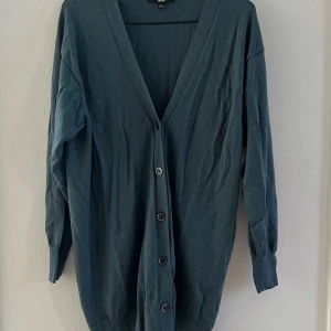 Mörkgrön kofta från Uniqlo - Säljer en tunnare mörkgrön/petroluem kofta från Uniqlo i 100% ull. Koftan har v-ringning, långa ärmar och knäppning framtill med fem knappar. Sparsamt använd, fint skick