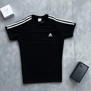 Svart t-shirt från Adidas med vita ränder - Svart t-shirt från Adidas med klassiska vita ränder på axlarna och litet Adidas-logga på bröstet. T-shirten har rund hals och är kortärmad, perfekt för sport eller vardag. Materialet är mjukt och andas bra.