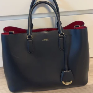 Svart handväska från Lauren Ralph Lauren - Stilren svart handväska från Lauren Ralph Lauren med röda detaljer på insidan och guldfärgade metalldetaljer. Väskan har dubbla handtag och en klassisk, rymlig form som passar perfekt för att bära med sig det viktigaste.