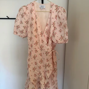 Ljusrosa blommig omlottklänning från & Other Stories - Supersöt ljusrosa omlottklänning från & Other Stories med små blommor i rosa och grönt. Klänningen har korta puffärmar, knyt i midjan och volangdetaljer längs nederkanten. Perfekt för vår och sommar!