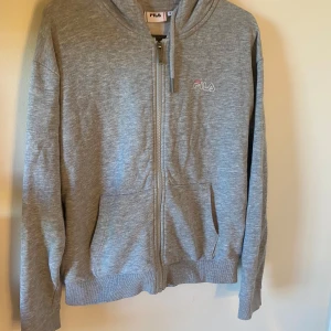 Grå hoodie från FILA - Säljer en klassisk grå hoodie från FILA med dragkedja framtill och två fickor. Tröjan har huva med snörning och diskret FILA-logga på bröstet. Perfekt för en avslappnad stil. Inga tecken på användning 