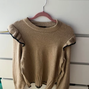 Beige stickad tröja med volang från Pernilla Wahlgren - Säljer en beige stickad tröja från Pernilla Wahlgren i storlek XS. Tröjan har volangdetaljer på axlarna med svarta kanter. Perfekt för kyligare dagar! Knappt använd 
