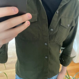 Mörkgrön skjorta/overshirt - Säljer en mörkgrön skjorta med knappar framtill och två bröstfickor med lock. Skjortan har lång ärm och klassisk krage, perfekt för en stilren look. Passar till jeans eller chinos.