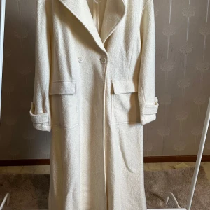 Vit kappa från NA-KD - Säljer en lång, vit oversized kappa från NA-KD (Hanna Schönberg pick) med klassisk krage och dubbelknäppning. Kappan har stora fickor framtill och uppvikta ärmslut. Helt oanvänd endast testat hemma och kunde inte returneras pga att jag drog av prislappen. Slutsåld online och kostade orginalpris 1299kr.