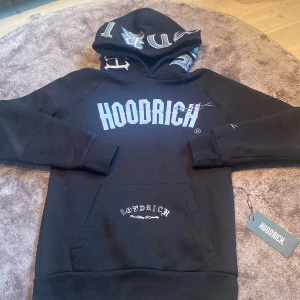Svart hoodie från Hoodrich - Säljer en svart hoodie från Hoodrich med stor vit logga på bröstet och tryck på både ficka och rygg. Hoodien har huva med text, magficka och ribbade muddar. Perfekt för en streetwear-look. Tag medföljer vid köp(använd typ 5ggr).                            Riktigt fint skick!                                                            Hör av dig vid minsta lilla!