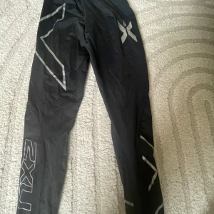 Svarta träningstights från 2XU - Svarta träningstights från 2XU med gråa reflexdetaljer och loggor längs benen. Sitter tight och är perfekta för löpning eller gym. Snygg sportig look med hög komfort och bra stöd. Etiketten är av klippt, osäker på storleken men gissar på xs/s