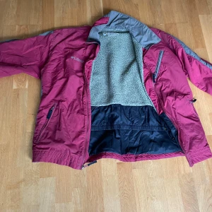Vinröd och grå vindjacka från Columbia - Säljer en vinröd och grå vindjacka från Columbia med dragkedja och flera fickor. Jackan har lång ärm och är fodrad med mjuk, ljusgrön fleece på insidan. Perfekt för kyliga dagar och har en sportig look.med byxor som hör till.Vääldigt bra för vintern eftersom den är väldigt varm och skön.