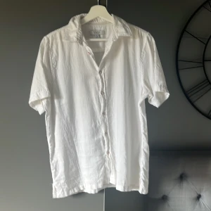 Vit kortärmad skjorta från SMOG - Säljer en vit kortärmad skjorta från SMOG i relaxed fit. Skjortan har klassisk krage och knäppning framtill. Perfekt för varma dagar och enkel att matcha med olika outfits.
