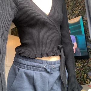 Svart ribbad omlottopp - Säljer en svart  omlottopp med långa ärmar och volangkant nertill. Tröjan är ribbstickad och har knyt i midjan för en snygg passform. 