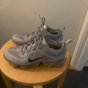 Grå Nike Air Vapormax sneakers - Säljer ett par grå Nike Air Vapormax sneakers med svart swoosh på sidan och transparent sula. Skorna har snörning och en sportig design med detaljer i orange. Perfekta för dig som gillar streetwear och komfort.