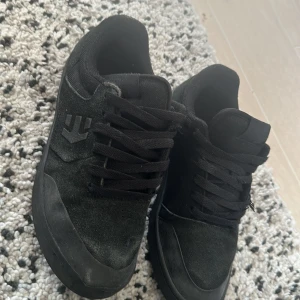 Svarta sneakers från Etnies - Säljer ett par svarta sneakers från Etnies med mockadetaljer och snörning. Skorna har rund tå och platt sula, perfekt för en avslappnad stil. Klassisk Etnies-logga på sidan och helsvart design.