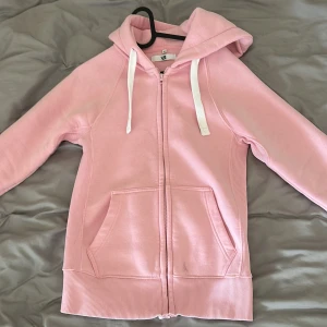Ljusrosa hoodie från Russedress - Säljer en ljusrosa hoodie från Russedress i storlek XS. Tröjan har dragkedja, huva med vita snören och två fickor framtill. Mjuk fleece på insidan och ribbade muddar vid ärmslut och nederkant. Perfekt för mysiga dagar. Endast använd en par gånger.