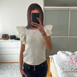 Vit ribbad topp med volangärmar från Zara - Säljer en vit ribbad topp från Zara med stora volangärmar. Toppen har rund halsringning och är tillverkad i ett mjukt material som sitter bekvämt. St S men passar perfekt även som en Xs! OBS finns inte att köpa längre!