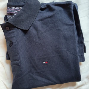 Mörkblå pikétröja från Tommy Hilfiger - Säljer en klassisk mörkblå pikétröja från Tommy Hilfiger med liten broderad logga på bröstet. Tröjan har krage, knappar framtill och är tillverkad i bomull. Perfekt för en stilren och avslappnad look.