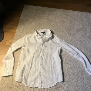 Vit linneskjorta från Polo Ralph Lauren - Klassisk vit skjorta i linne från Polo Ralph Lauren med blå broderad logga på bröstet. XL i barnstorlek motsvarar XS/S i vuxenstorlek. Skjortan har lång ärm, knappar framtill och en avslappnad passform. Perfekt för en stilren och fräsch look.