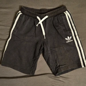 Svarta shorts från Adidas med vita ränder - Svarta Adidas shorts med vita sidoremsor och klassisk Adidas-logga på vänster ben. Dragsko i midjan och bekväm passform. Perfekta för träning eller chill. Materialet känns mjukt och sportigt.