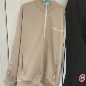 Beige track jacket från Palm Angels - Säljer en beige track jacket från Palm Angels med vit dragkedja och logga på bröstet. Jackan har hög krage, långa ärmar och två fickor med dragkedja. Perfekt för en sportig och trendig look. Anväd max 5 gånger super fin