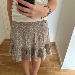 Beige blommig volangkjol från 157 - Supersöt kjol från lager 157 i beige med vitt blommönster. Kjolen är väldigt stretchig och har volang nertill som ger en fin och luftig känsla. Perfekt för varma dagar och enkel att matcha med en enkel topp.