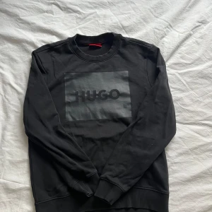 Svart sweatshirt från Hugo Boss - Säljer en svart sweatshirt från Hugo Boss med stor logga framtill i mörkgrått. Tröjan har rund halsringning, långa ärmar och ribbade muddar vid ärmslut och nederkant. Perfekt för en stilren och enkel look. Passar också till S använd få gånger 