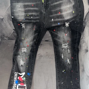 Svarta jeans från Dsquared2 med färgstänk - Säljer ett par svarta jeans från Dsquared2 med slitningar, färgstänk i flera färger och tryck på ena benet. Jeansen har normal passform och klassisk femficksdesign. Snygga detaljer som logga och slitna partier.