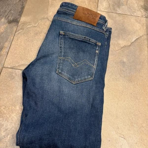 Blå jeansbyxor från Replay - Säljer ett par klassiska blå jeans från Replay med femficksdesign och läderpatch bak i midjan. Jeansen har raka ben och normal passform, tillverkade i slitstarkt denimtyg. Perfekta för en avslappnad stil.