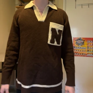 Brun vintageinspirerad stickad tröja med krage - Retrobrun stickad tröja med vit krage och stor vit ficka med bokstaven N på bröstet. Tröjan har långa ärmar och vita detaljer vid midjan och halsen. Perfekt för dig som gillar unik och klassisk stil.