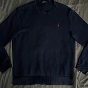Mörkblå stickad tröja från Polo Ralph Lauren - Säljer en klassisk mörkblå stickad tröja från Polo Ralph Lauren med rund halsringning och den ikoniska röda loggan broderad på bröstet. Tröjan har långa ärmar och ribbade muddar vid ärmslut och nederkant. Perfekt för lager-på-lager-stil.