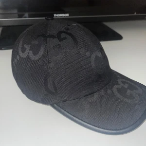 Gucci keps - Säljer en svart keps från Gucci med GG-mönster. Kepsen har justerbart svart bälte baktill i skinn och är tillverkad i bomullsmaterial. Perfekt accessoar för en trendig look.