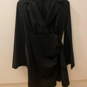 Svart långärmad klänning  - Säljer en svart kortklänning med lång ärm och omlottmodell. Klänningen har kavajkrage. Perfekt för dig som gillar stilrena och eleganta plagg.
