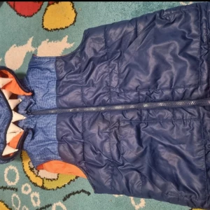 Blå dunväst med monsterhuva H&M - Säljer en blå dunväst från H&M med coolt monstertryck på huvan, vita mjuka tänder och stora ögon. Västen har orange foder, dragkedja framtill och två fickor. Perfekt för dig som vill sticka ut med en lekfull och unik look.