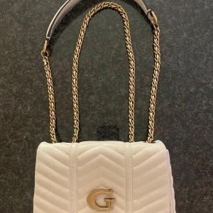 Vit quiltad axelväska från Guess - Snygg vit axelväska från Guess med quiltad design och guldfärgad kedjerem. Väskan har ett stort G i metall framtill och flera fack inuti för kort och småsaker. Perfekt storlek för det viktigaste och riktigt trendig look. Créme vit i färgen. 