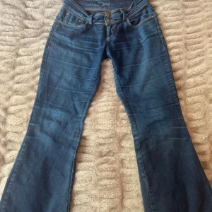 Blå bootcut jeans med låg midja - Snygga blå bootcut jeans från Jane Norman med dubbla knappar i midjan och klassiska fickor. Jeansen har en lägra midja. Liten slitning innanför se bild. Fråga om mer mått om det behövs