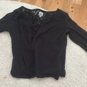 Svart kofta med spetsdetaljer från Time - Säljer en svart kofta från Time med fina spetsdetaljer upptill vid axlar och hals. Koftan har långa ärmar och en klassisk passform, perfekt att slänga över en topp för en snygg look.