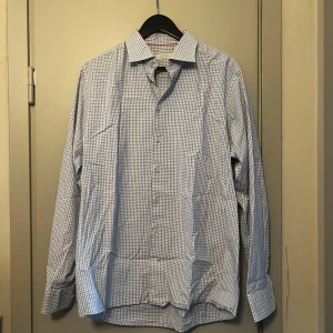 Rutig skjorta från Eton - Säljer en klassisk långärmad skjorta från Eton i blått och vitt rutigt mönster. Skjortan har knappar framtill och är tillverkad i bomull. Perfekt för dig som gillar stilrena och tidlösa plagg.