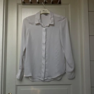 Vit blus från H&M - Klassisk vit blus från H&M med lång ärm och krage. Skjortan har en enkel och stilren design med knappar framtill och är tillverkad i ett lätt och mjukt material. Perfekt för en clean och tidlös look.