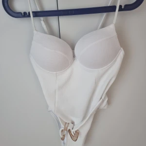 Vit baddräkt med bälte - Säljer en snygg vit baddräkt med vadderade kupor och justerbara axelband. Baddräkten har ett dekorativt bälte med guldfärgat spänne i midjan och är tillverkad i ett ribbat material. Perfekt för stranden eller poolen! Oanvänt 