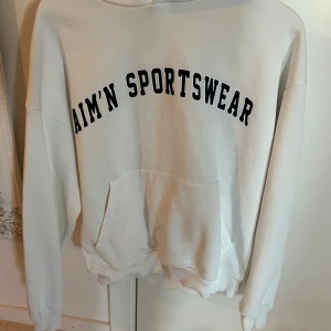 Vit hoodie från Aim'n Sportswear - Säljer en vit hoodie från Aim'n Sportswear med svart tryck framtill. Tröjan har huva, magficka och ribbade muddar. Tillverkad i en mjuk bomulls- och polyestermix, perfekt för en avslappnad stil. Använd endast en gång. 