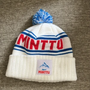 Minttu mössa  - Säljer en vit stickad mössa från Minttu med blå ränder och röd text. Mössan har en blåvit tofs på toppen och en uppvikt kant med Minttu-logga framtill. Perfekt för kalla dagar och snygg detalj till vinterlooken.