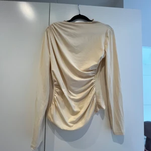 Beige långärmad topp med drapering - Säljer en beige långärmad topp med snygg drapering på sidan. Toppen har en mjuk och glansig yta och är figurnära i modellen. Perfekt för dig som vill ha något stilrent men ändå med en twist.