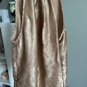 Champagnefärgad ärmlös topp i glansigt material - Säljer en snygg champagnefärgad topp med halterneck och ärmlös design. har ett glansigt, satinliknande material som ger en lyxig känsla. Perfekt för dig som vill sticka ut med en elegant look.