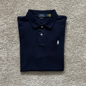 Mörkblå pikétröja från Polo Ralph Lauren - Säljer en klassisk mörkblå pikétröja från Polo Ralph Lauren med vit broderad logga på bröstet. Tröjan är i nyskick och är näst intill oanvänd. Pris kan diskuteras vid snabb affär!