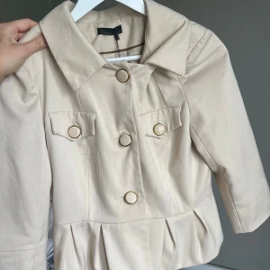 Beige kavajjacka från Vero Moda - Stilren beige kavajjacka från Vero Moda med markerad krage, dekorativa ficklock och stora guldiga knappar framtill. Jackan har veckad nederkant och trekvartsärmar för en trendig look.
