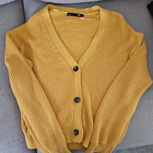 Gul stickad kofta med knappar - Säljer en gul stickad kofta från 157 med v-ringning och svarta knappar framtill. Koftan har långa ärmar och ribbade muddar vid ärmslut och nederkant. Perfekt att slänga på sig när det är lite kyligt ute!