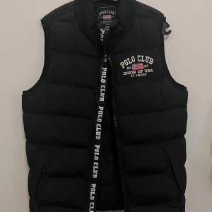 Svart dunväst från Polo Club - Svart dunväst från Polo Club med broderad logga och text på bröstet. Västen har dragkedja framtill och två fickor på sidorna. Perfekt för lager-på-lager och enkel att matcha med olika outfits.