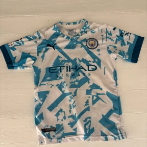 Manchester City bortatröja 2020/21 - Säljer en Manchester City bortatröja från säsongen 2020/21 med blått och vitt abstrakt mönster. Tröjan har korta ärmar, klubbmärke på bröstet och sponsortryck. Nummer och namntryck på ryggen. Perfekt för fotbollsfans!