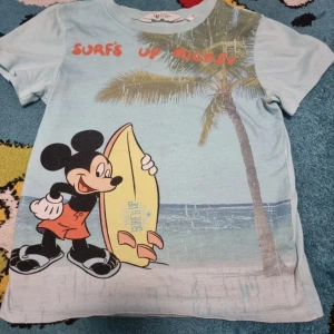 T- shirt med Mickey Mouse  - Säljer en ljusblå t-shirt med tryck av Mickey Mouse som håller i en gul surfbräda framför en palm och hav. Texten 'Surf's Up Mickey' i orange upptill. Perfekt för dig som gillar surf och Disney!