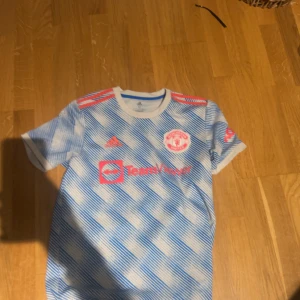 Manchester United bortatröja 2021/22 - Säljer en Manchester United bortatröja från säsongen 2021/22. Tröjan är kortärmad med blått och vitt randigt mönster, röda detaljer och Adidas-logga. Materialet är lätt och andas, perfekt för fotbollsträning eller match.